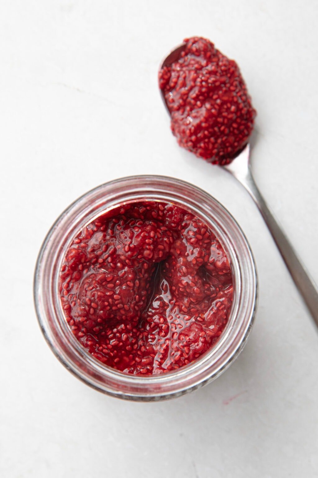A bird-eye view of a jar of raspberry chia jam, filled near the top. A spoon of chia jam lies next to it.
