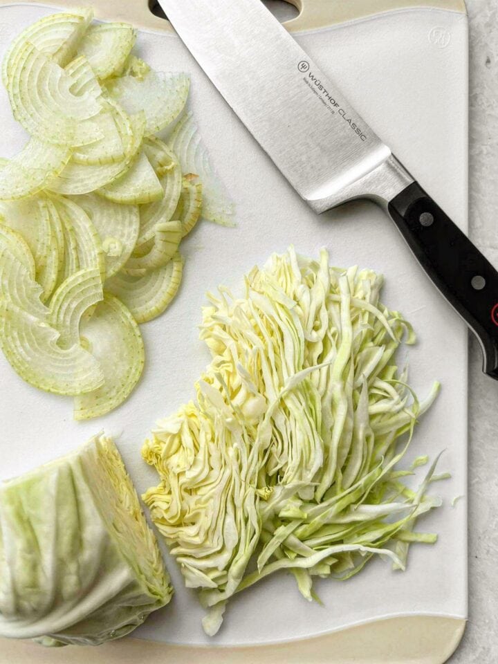 A white chopping board with half-moon onions, sliced cabbage and a chefs knife.