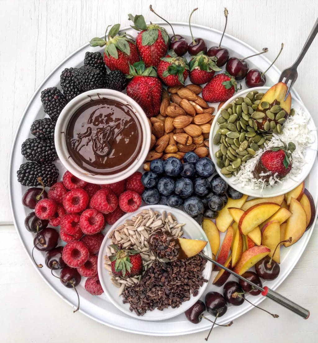 A fruit and dark chocolate fondue platter with berries, summer fruit, coconut and seeds.