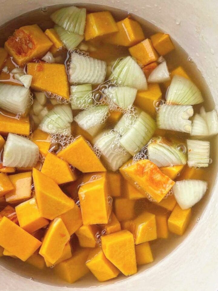 A soup pot with butternut squash cubes, diced onion and garlic, and vegetable stock, before being cooked.