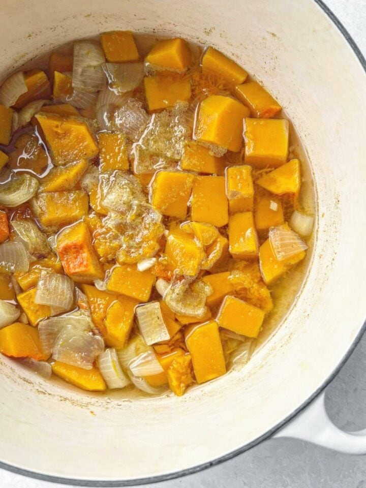 A soup pot with cooked butternut squash cubes, diced onion and garlic, and vegetable stock.