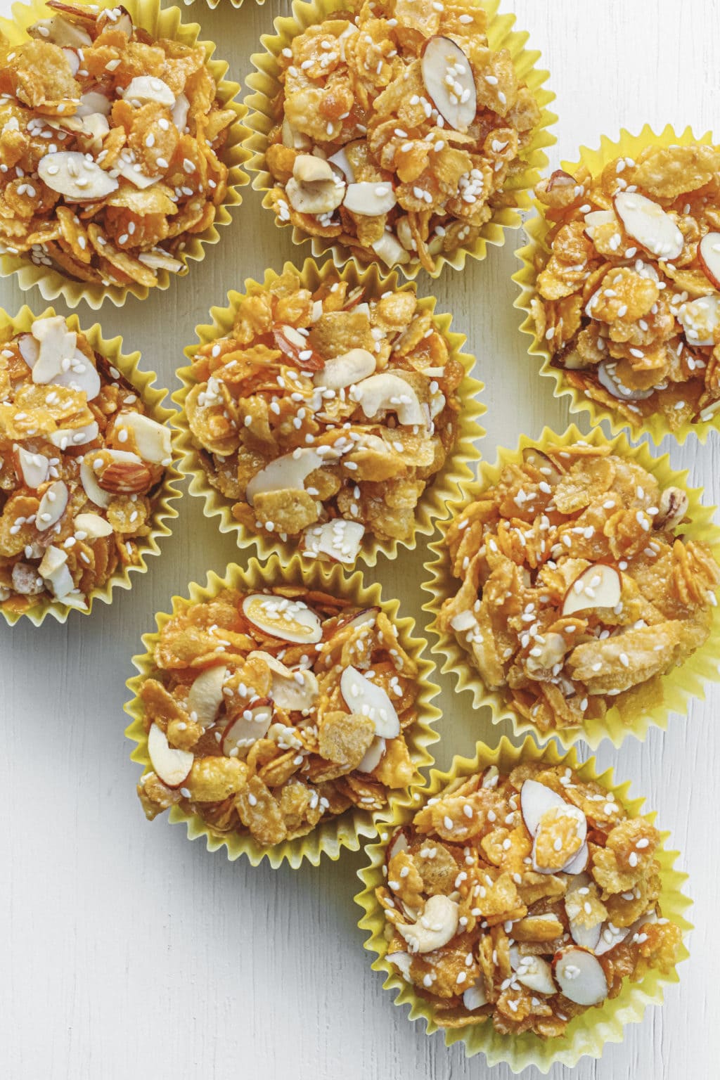 Honey joys on a white table. The honey joys are made with sliced almonds, sesame seeds, cornflakes and cashews.