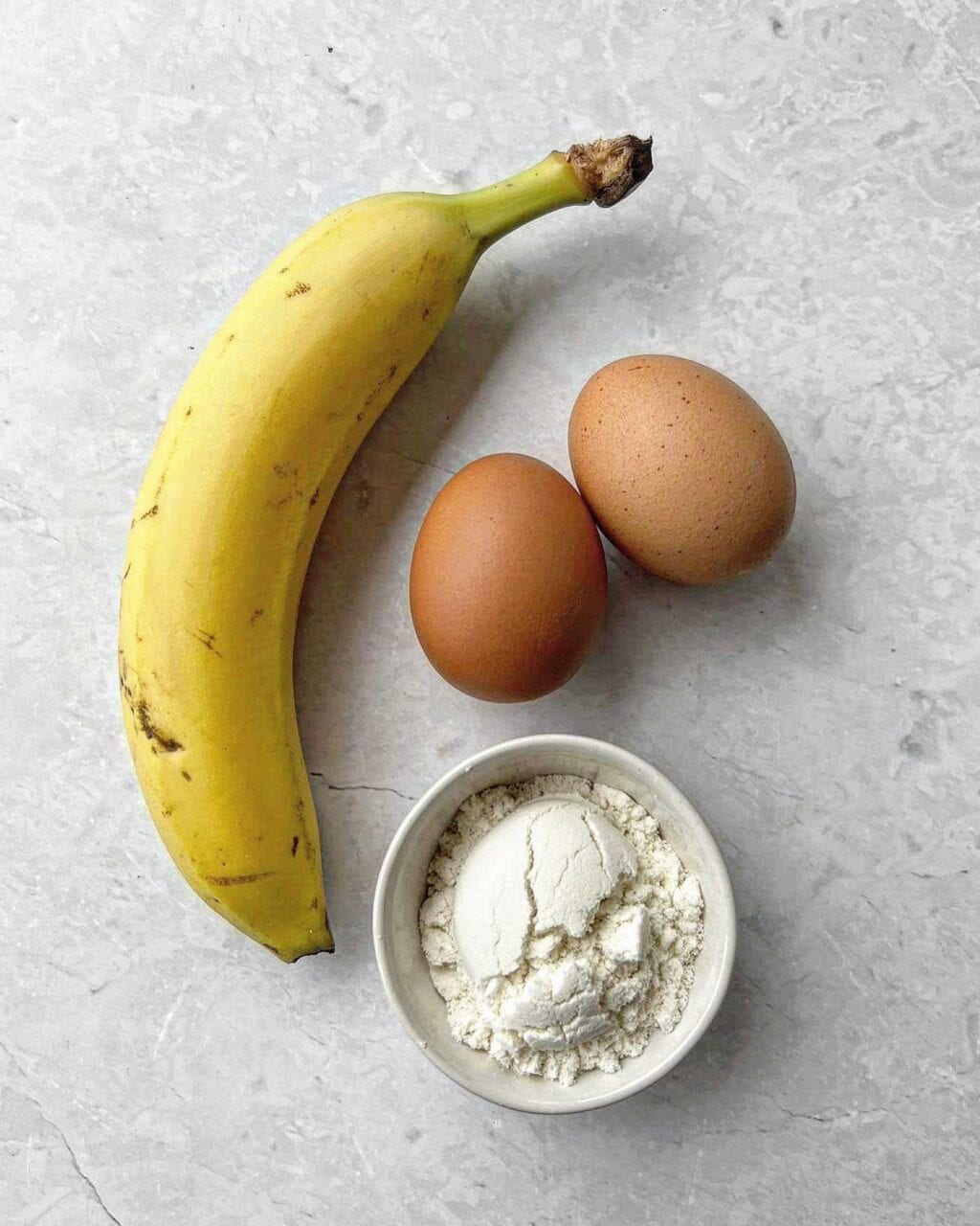 A photo of the ingredients needed to make banana protein pancakes, including a whole banana, a small bowl of protein powder and two eggs.