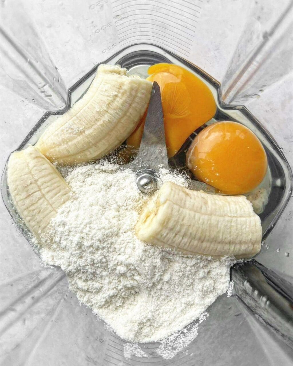 A photo of a banana broken into chunks, 2 eggs and protein powder in a blender.