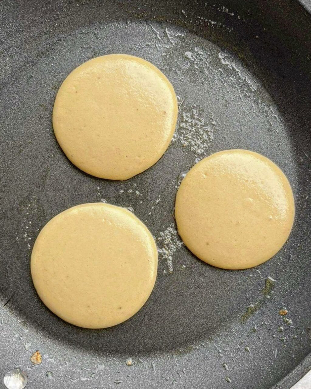 A photo of three batter rounds of protein pancakes in a black pan.