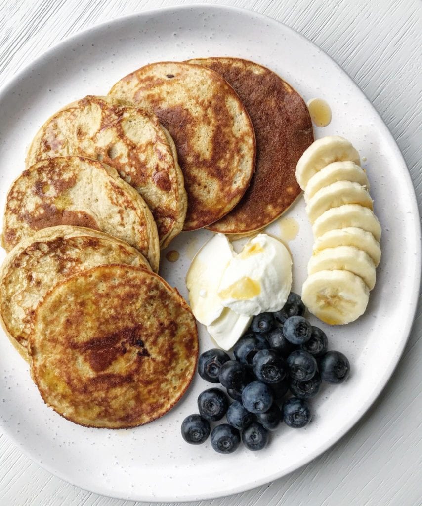 Protein pancakes served on a white plate, with sliced banana, yoghurt, blueberries and a drizzle of maple syrup. 