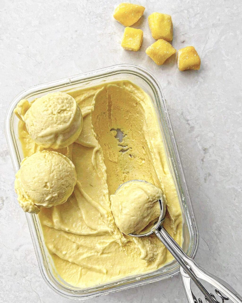 A birds-eye photo of mango coconut ice cream with pieces of mango to the side and a loaded ice cream scoop