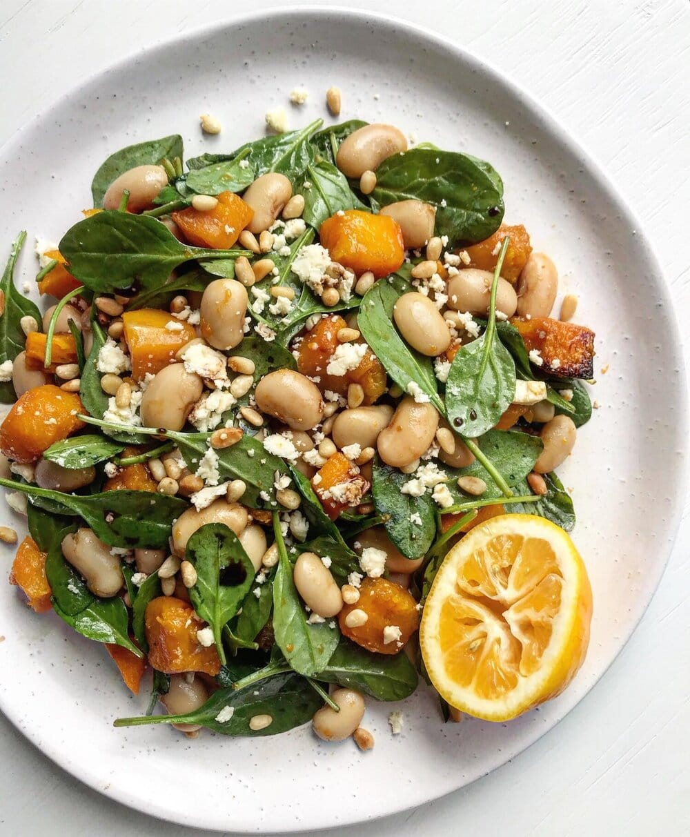 A close up of a roasted honey-pumpkin salad tossed wit spinach, white beans, feta and pine nuts. A half lemon is a garnish. 