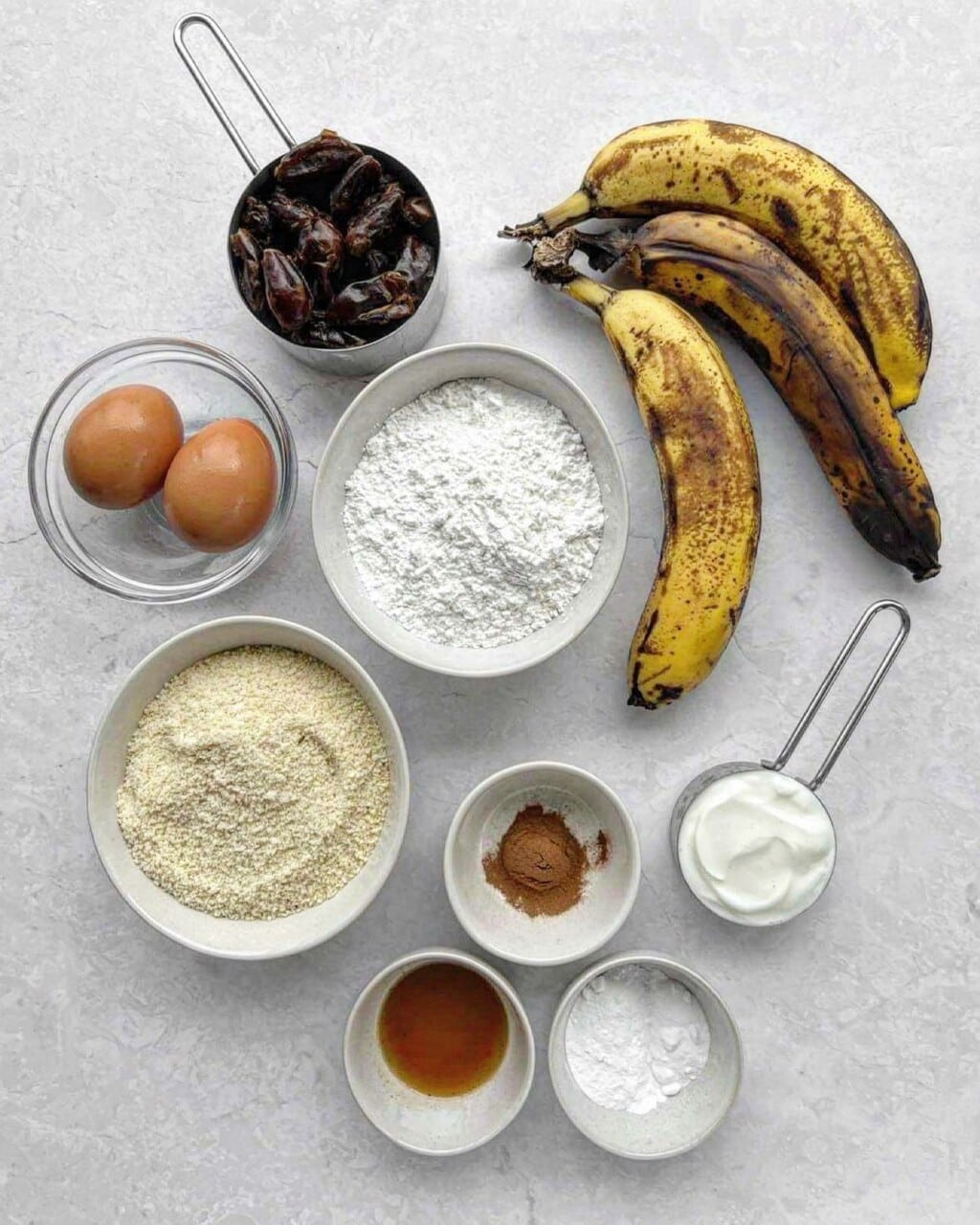 A birds-eye photo of the ingredients needed to make banana date loaf, including spotty yellow bananas, dates, eggs, flour, yoghurt, vanilla, cinnamon and greek yoghurt.