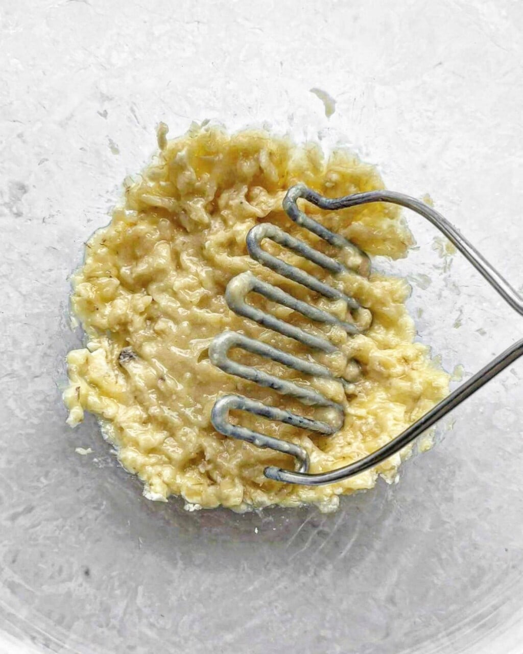 Banana being mashed in a bowl with a potato masher.