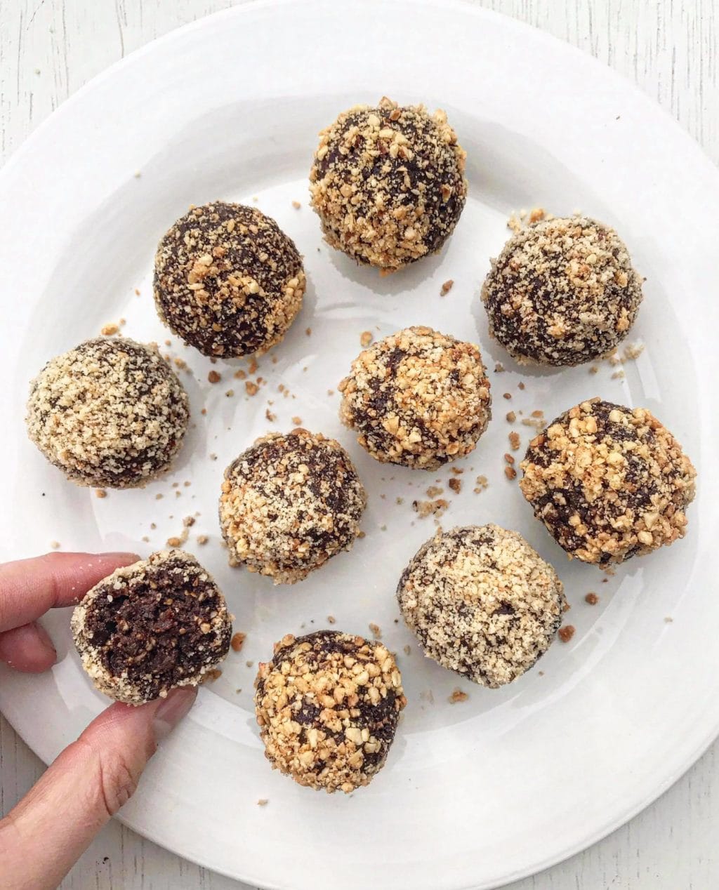 A picture of ten chocolate hazelnut bliss balls on a white plate. One is being held and is cut in half to see the inside.