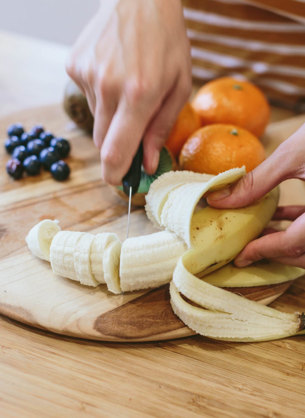 Banana being sliced by a knife on a chopping board, surrounded by fruit.