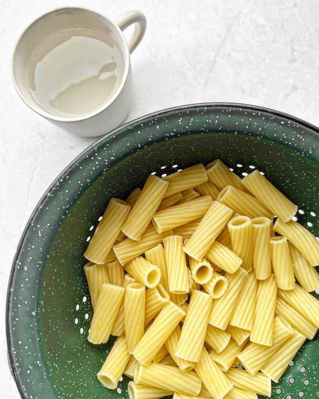 Cooked pasta in a colander, with a cup of reserved pasta water on the side.