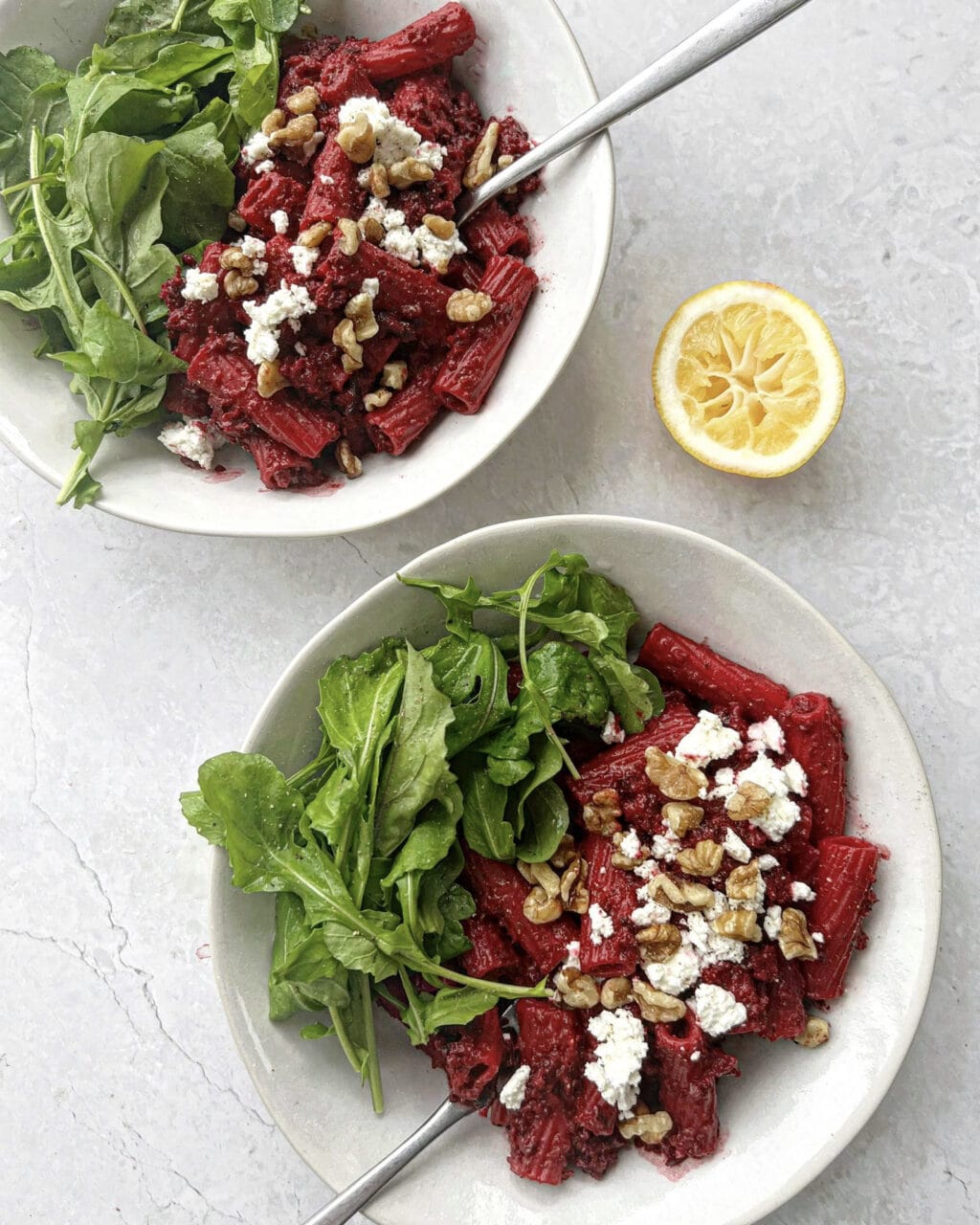 Two bowls of beetroot goat cheese pasta topped with goat cheese and chopped walnuts, with rocket on the side. A halved lemon is next to the plates.