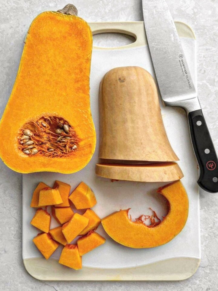A butternut squash being prepared for soup. The squash is cut in half, with one half left whole and one half being sliced into cubes and wedges. There is a chefs knife on the chopping board.