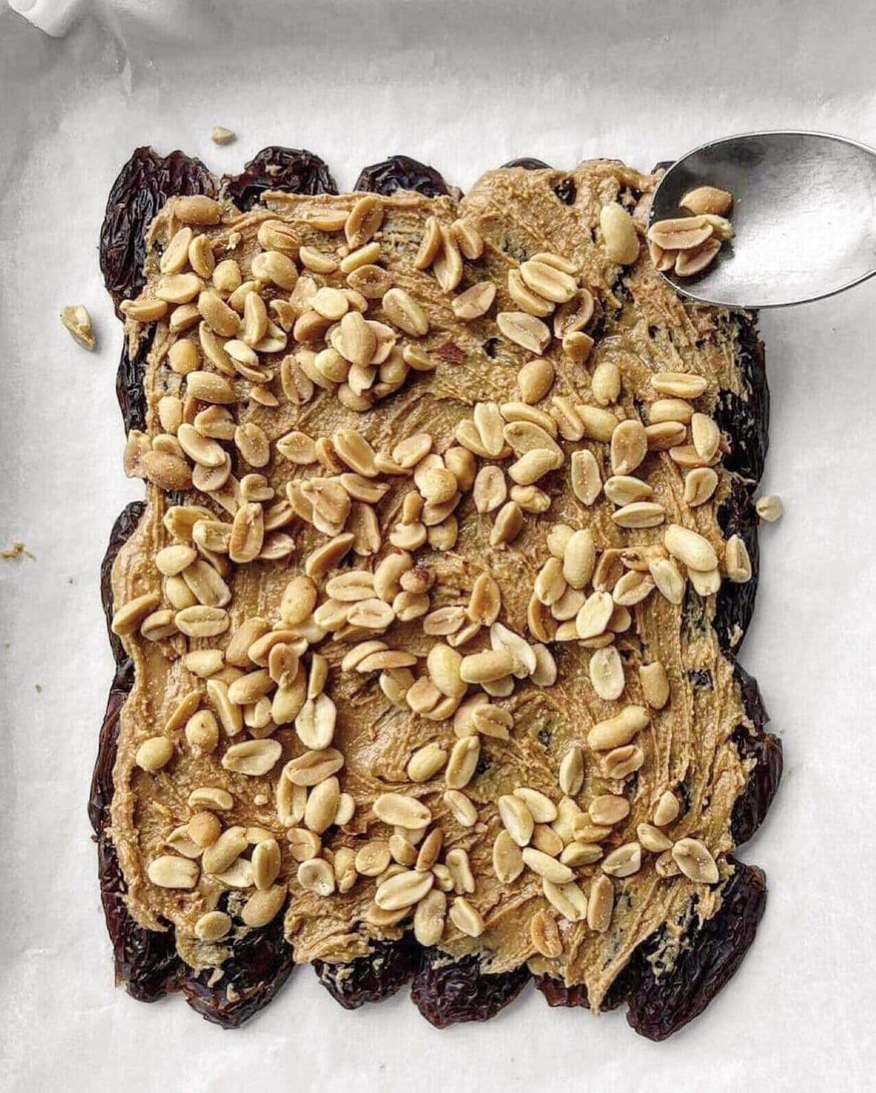 Roasted salted peanuts being sprinkled overtop peanut butter spread medjool dates on a line baking tray.