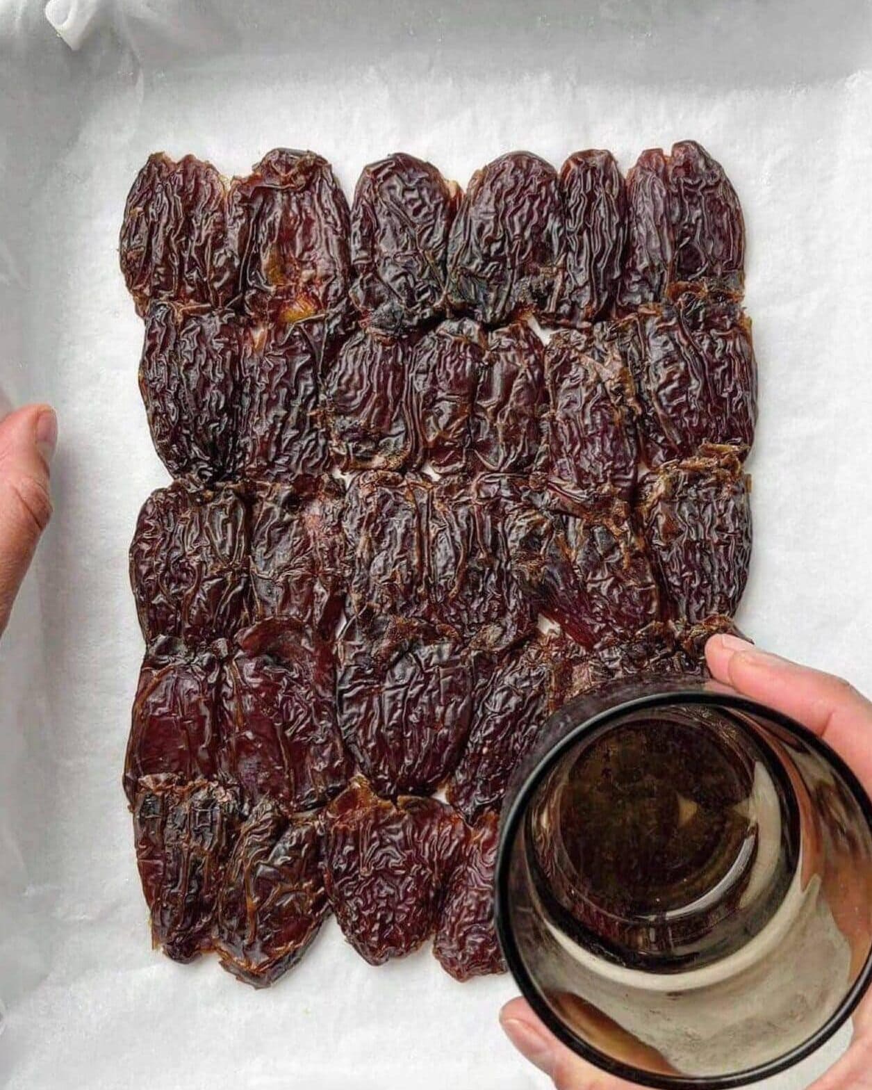 Medjool dates on a lined baking tray being pressed down flat with the bottom of a glass.