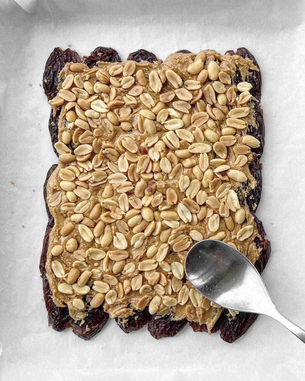 Salted roasted peanuts being pressed down with the back of a spoon onto  flattened medjool dates that have been spread with peanut butter.