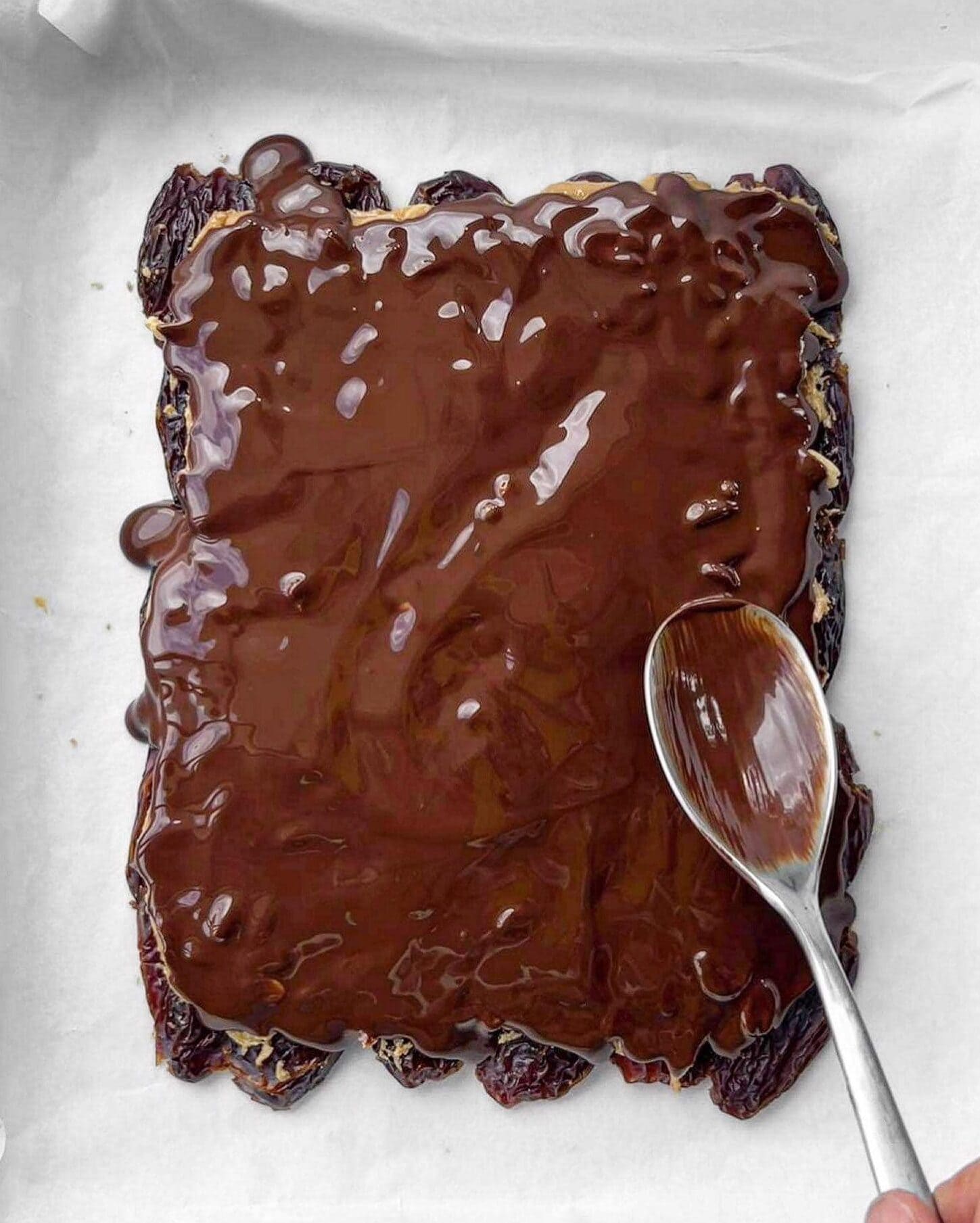 Flattened medjool dates being covered and spread with melted dark chocolate using the back of a spoon.